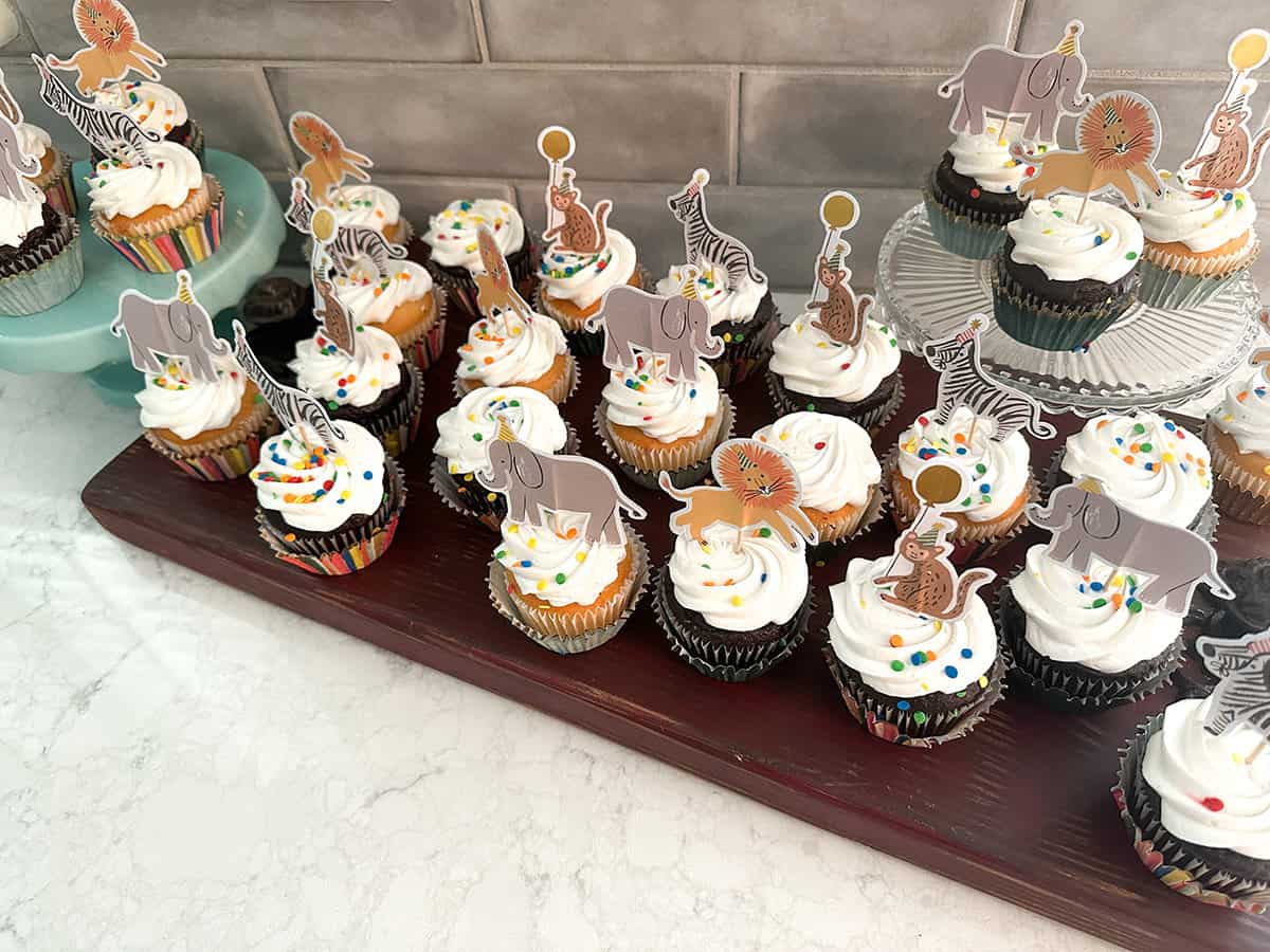 cupcakes with colorful animal toppers wearing birthday hats set on large platters ready for an animal themed second birthday party