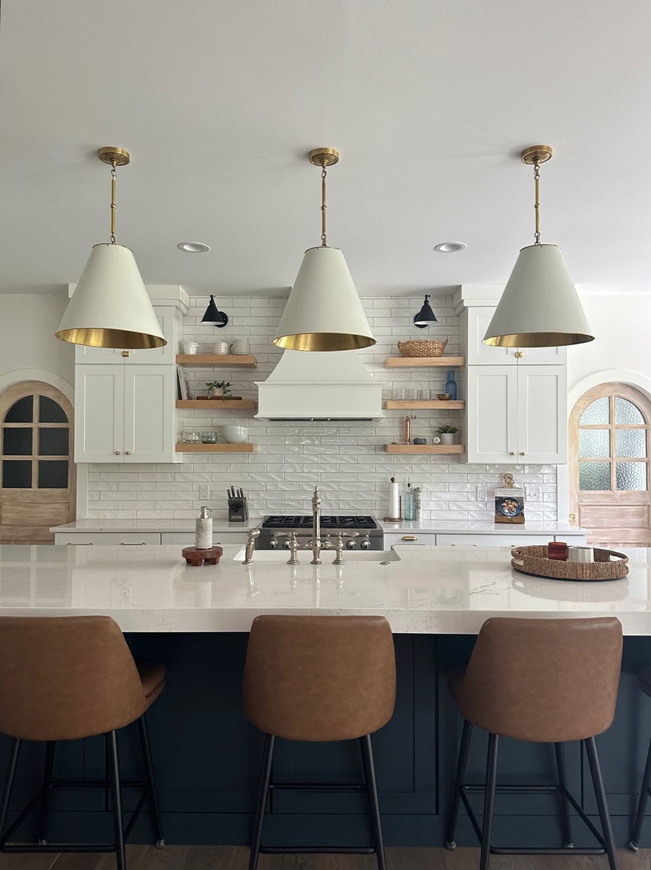 white transitional kitchen with white large pendants that have a brass interior hanging over kitchen island that has white quartz counters and floating shelves behind on a range wall