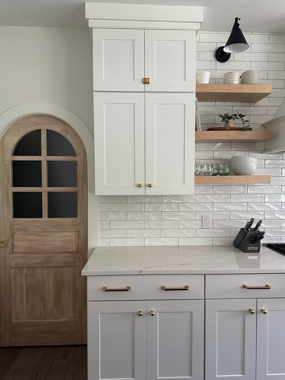 White kitchen cabinets and white backsplash with wood floating shelves, black sconces, and wood arched doors on either side