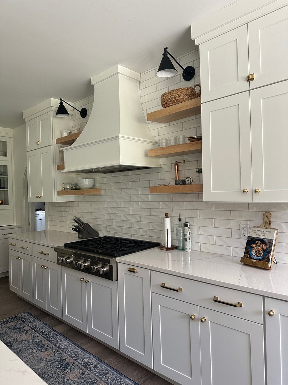 White kitchen cabinets and backsplash with wood floating shelves and black sconces hung looking down over a quartz countertop