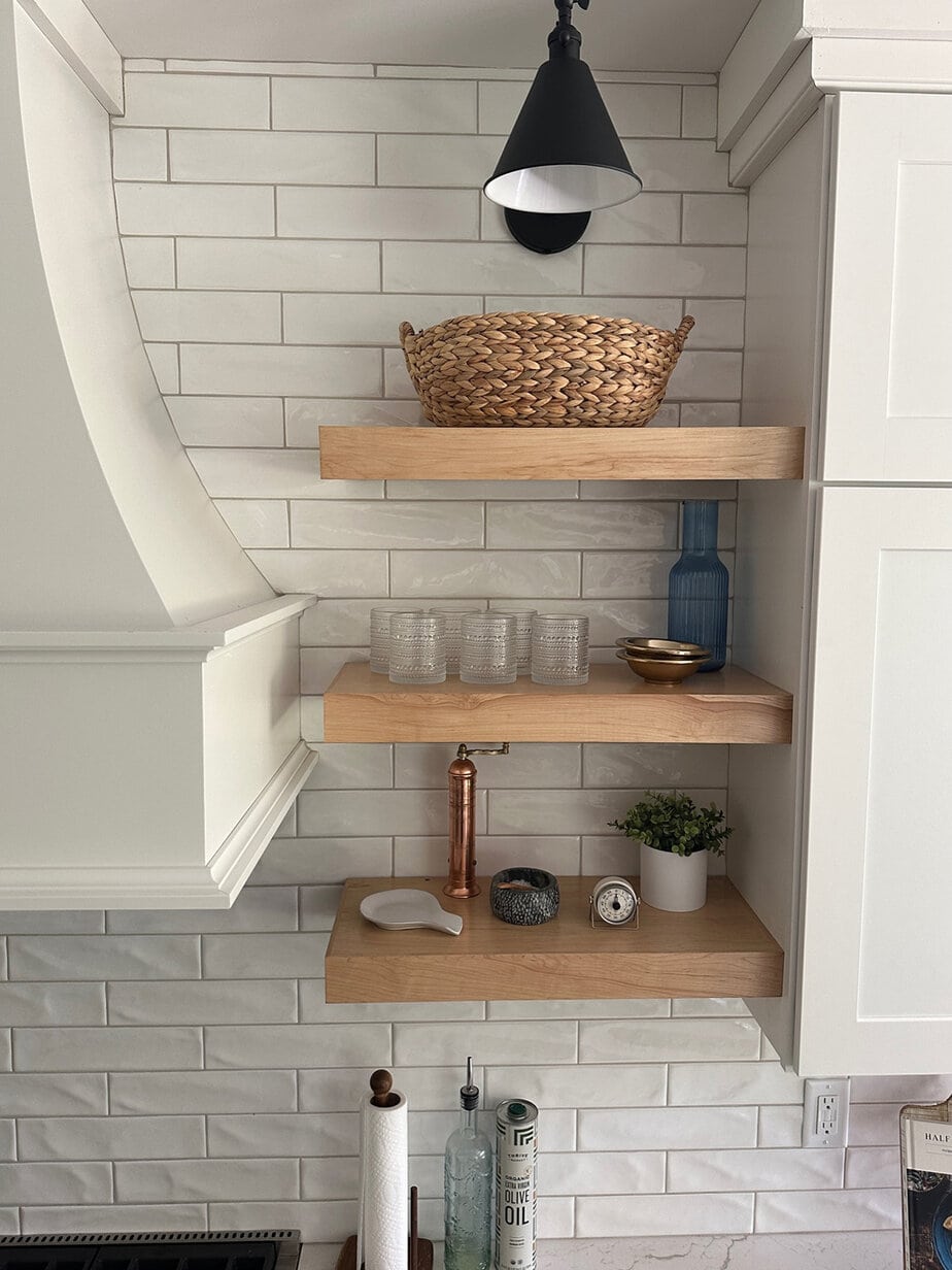 White kitchen cabinets and white backsplash with wood floating shelves, black sconces, and wood arched doors on either side